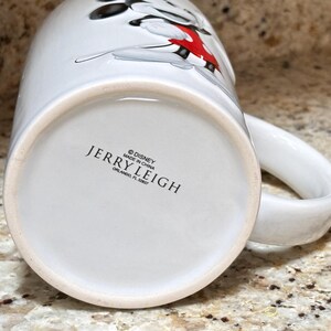 K&ouml;nnte beinhalten: Wei&szlig;er Keramikbecher mit einem Cartoon-Charakter-Design in Schwarz, Rot und Wei&szlig;. Der Becher hat einen gro&szlig;en Henkel und den Text "JERRY LEIGH" und "&copy; DISNEY MADE IN CHINA ORLANDO, FL 32837" auf der Unterseite.