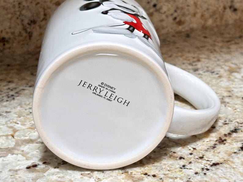 K&ouml;nnte beinhalten: Wei&szlig;er Keramikbecher mit einem Cartoon-Charakter-Design in Schwarz, Rot und Wei&szlig;. Der Becher hat einen gro&szlig;en Henkel und den Text "JERRY LEIGH" und "&copy; DISNEY MADE IN CHINA ORLANDO, FL 32837" auf der Unterseite.