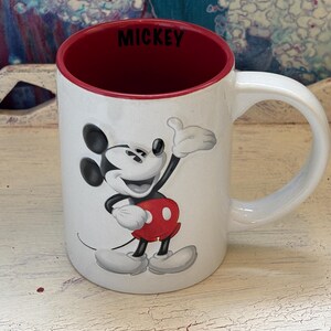 K&ouml;nnte beinhalten: Wei&szlig;er Keramikbecher mit Mickey Mouse, rotem Innenraum und Griff. Der Becher hat ein Cartoon-Bild von Mickey Mouse, die einen Daumen hoch gibt. Das Wort "MICKEY" ist innen schwarz gedruckt.