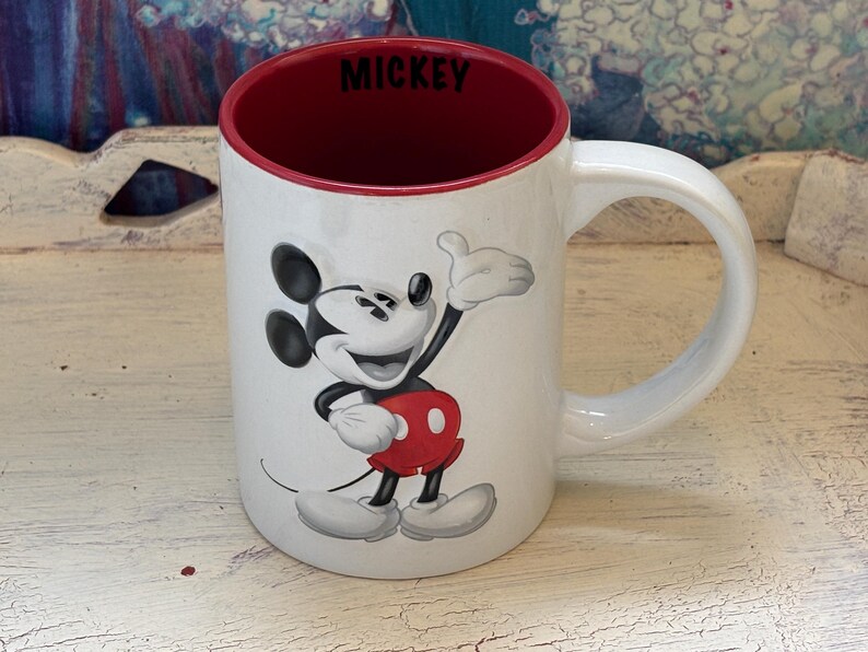 K&ouml;nnte beinhalten: Wei&szlig;er Keramikbecher mit Mickey Mouse, rotem Innenraum und Griff. Der Becher hat ein Cartoon-Bild von Mickey Mouse, die einen Daumen hoch gibt. Das Wort "MICKEY" ist innen schwarz gedruckt.