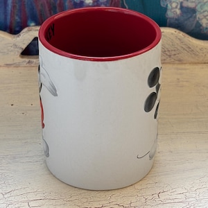 K&ouml;nnte beinhalten: Wei&szlig;er Keramikbecher mit rotem Innenraum und Rand. Der Becher hat ein Cartoon-Charakter-Design mit schwarzen und grauen Akzenten. Der Becher ist zylindrisch und steht auf einer wei&szlig;en, abgenutzten Oberfl&auml;che.