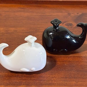 May include: A pair of ceramic whale-shaped salt and pepper shakers. One shaker is glossy black, and the other is white. Both have a tail fin, a spout on top, and small side fins. The shakers are on a wooden surface.