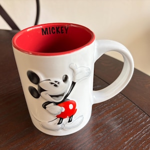 K&ouml;nnte beinhalten: Wei&szlig;e Keramiktasse mit einem erhabenen Mickey Mouse-Motiv in Schwarz, Rot und Wei&szlig;. Das Innere der Tasse ist rot, mit dem Wort "MICKEY" in Schwarz aufgedruckt. Sie hat einen gro&szlig;en, gebogenen Griff.