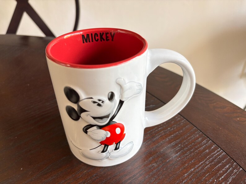 K&ouml;nnte beinhalten: Wei&szlig;e Keramiktasse mit einem erhabenen Mickey Mouse-Motiv in Schwarz, Rot und Wei&szlig;. Das Innere der Tasse ist rot, mit dem Wort "MICKEY" in Schwarz aufgedruckt. Sie hat einen gro&szlig;en, gebogenen Griff.