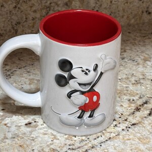 K&ouml;nnte beinhalten: Wei&szlig;er Keramikbecher mit roter Innenseite und wei&szlig;em Henkel. Der Becher zeigt ein erhabenes Bild von Mickey Mouse in seinem klassischen Outfit, mit schwarzen Ohren, roter Hose und wei&szlig;en Handschuhen. Der Becher steht auf einer gesprenkelten Oberfl&auml;che.