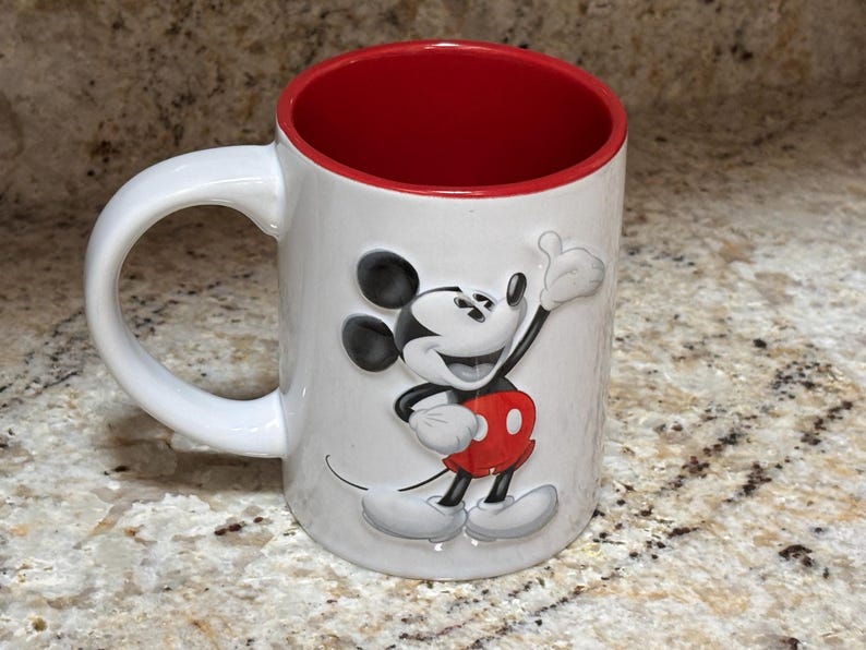 K&ouml;nnte beinhalten: Wei&szlig;er Keramikbecher mit roter Innenseite und wei&szlig;em Henkel. Der Becher zeigt ein erhabenes Bild von Mickey Mouse in seinem klassischen Outfit, mit schwarzen Ohren, roter Hose und wei&szlig;en Handschuhen. Der Becher steht auf einer gesprenkelten Oberfl&auml;che.