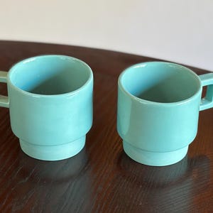 May include: Two matching turquoise ceramic mugs with square handles. The mugs have a stacked design, with a wider base and a narrower top. The mugs are sitting on a dark brown wooden surface.