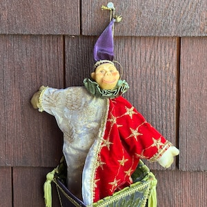 May include: A decorative jester doll with a smiling face, wearing a purple hat and a red and gold star-patterned coat. The doll is in a green and gold basket, hanging against a brown wooden background.