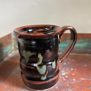 May include: A cylindrical ceramic mug with a glossy finish, featuring a dark black and deep brown glaze. The mug has a wide, flared rim and a curved handle, with subtle, irregular patches of light blue and grey color accents along the body.