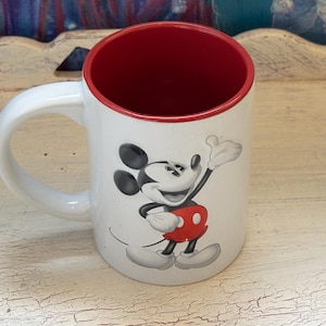 K&ouml;nnte beinhalten: Wei&szlig;er Keramikbecher mit roter Innenseite und wei&szlig;em Henkel. Der Becher zeigt eine l&auml;chelnde Mickey Mouse-Grafik in Schwarz, Rot und Wei&szlig;. Ein Disney-Klassiker.