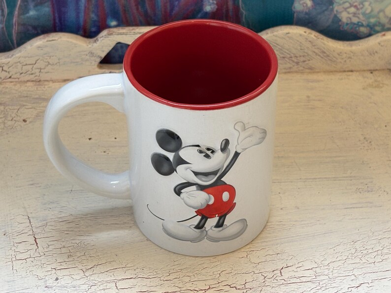 K&ouml;nnte beinhalten: Wei&szlig;er Keramikbecher mit roter Innenseite und wei&szlig;em Henkel. Der Becher zeigt eine l&auml;chelnde Mickey Mouse-Grafik in Schwarz, Rot und Wei&szlig;. Ein Disney-Klassiker.