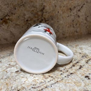 K&ouml;nnte beinhalten: Wei&szlig;er Keramikbecher mit Henkel, mit einem Design mit roten und schwarzen Akzenten. Auf dem Boden der Tasse steht der Text "&copy; Disney JERRY LEIGH MADE IN CHINA". Der Becher steht auf einer gesprenkelten Oberfl&auml;che.