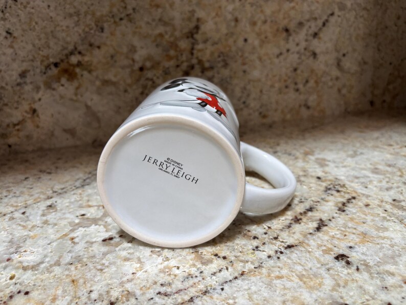 K&ouml;nnte beinhalten: Wei&szlig;er Keramikbecher mit Henkel, mit einem Design mit roten und schwarzen Akzenten. Auf dem Boden der Tasse steht der Text "&copy; Disney JERRY LEIGH MADE IN CHINA". Der Becher steht auf einer gesprenkelten Oberfl&auml;che.