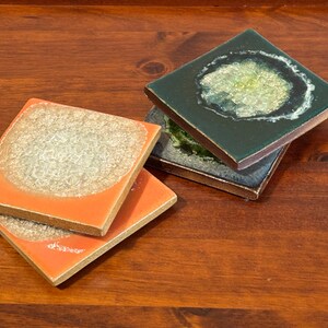May include: Three square coasters with a glossy finish. Two coasters are orange with a textured, light-colored circular center. The third coaster is dark green with a textured, light-colored circular center and is stacked on a dark gray coaster.