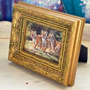 May include: A small, rectangular gold-colored picture frame with an ornate, detailed design. The framed artwork depicts three tiger cubs in a natural setting. The frame is propped up on a black stand.