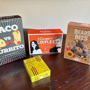 Może przedstawiać: Cztery pudełka z grami planszowymi są wyświetlane na drewnianej powierzchni. Gry obejmują "Taco vs Burrito", "Celebrity Couples", "Bears & Bees" oraz ż&oacute;łte pudełko z napisem "Taco Cat Cheese Pizza". Pudełka są kolorowe i zawierają ilustracje.