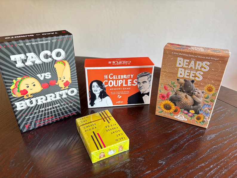 Może przedstawiać: Cztery pudełka z grami planszowymi są wyświetlane na drewnianej powierzchni. Gry obejmują "Taco vs Burrito", "Celebrity Couples", "Bears & Bees" oraz ż&oacute;łte pudełko z napisem "Taco Cat Cheese Pizza". Pudełka są kolorowe i zawierają ilustracje.