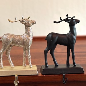 May include: Two deer-shaped stocking holders, one with a silver glitter finish on a light gold base and the other with a matte black finish on a matching black base. Each deer figure features metallic antlers and a hook extending from the bottom.