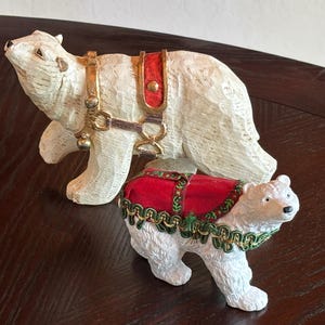 May include: Two white polar bear figurines. One bear is larger and has a red and gold harness. The smaller bear has a red saddle with green and gold trim. Both are on a dark brown surface.