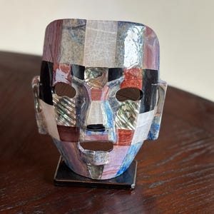 May include: A mosaic-style sculpture of a human face. The face is composed of various rectangular pieces in shades of blue, red, white, black, and iridescent materials. The sculpture has a square base and is displayed on a dark wooden surface.