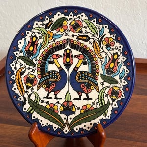 May include: Decorative ceramic plate with a vibrant peacock design. The plate features a dark blue rim and a white center with colorful floral and bird motifs. The plate is displayed on a wooden stand.