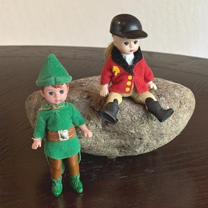 May include: Two small dolls, one dressed in a green tunic, pointed hat, brown belt, and green shoes, and the other wearing a red jacket with a black collar, yellow buttons, tan pants, black boots, and a black riding helmet, both with painted faces.