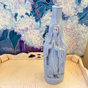 May include: A light blue ceramic vase with a detailed relief of a religious figure in prayer. The vase is displayed on a distressed white tray, with a colorful abstract painting in the background. The vase is approximately 10 inches tall.