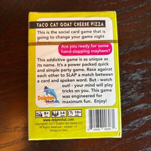 Może przedstawiać: Ż&oacute;łte pudełko do gry karcianej "Taco Cat Goat Cheese Pizza" z tekstem "Czy jesteś gotowy na chaos klaps&oacute;w?" Pudełko zawiera r&oacute;wnież logo delfina i kod kreskowy.