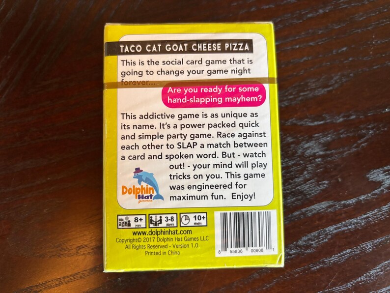 Może przedstawiać: Ż&oacute;łte pudełko do gry karcianej "Taco Cat Goat Cheese Pizza" z tekstem "Czy jesteś gotowy na chaos klaps&oacute;w?" Pudełko zawiera r&oacute;wnież logo delfina i kod kreskowy.