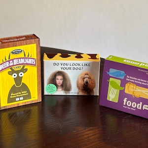 May include: Three board game boxes on a dark wooden surface. The games are "Deer in Headlights Game", "Do You Look Like Your Dog? A Memory Game", and "Food Smarts". The "Deer in Headlights" box has a deer graphic. The "Food Smarts" box is purple and green.