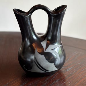 May include: A black pottery vase with a distinctive double-spout design and a handle. The vase features a stylized, light gray floral pattern on its rounded body. The vase is displayed on a dark wooden surface.