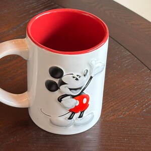 K&ouml;nnte beinhalten: Wei&szlig;er Keramikbecher mit roter Innenseite und gro&szlig;em Henkel. Der Becher zeigt ein erhabenes Bild von Mickey Mouse in seinem klassischen Outfit, mit schwarzen Ohren, roten Shorts und wei&szlig;en Handschuhen. Der Becher steht auf einer dunklen Holzoberfl&auml;che.