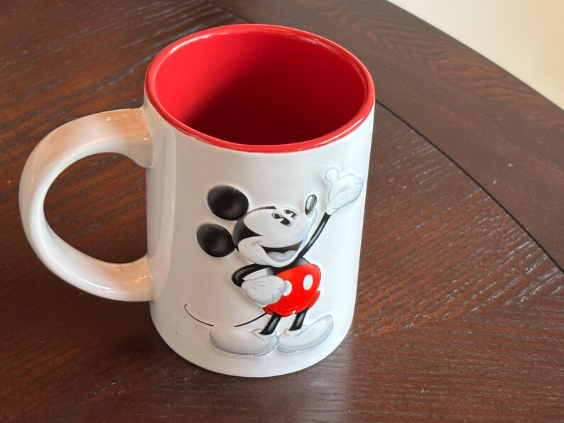 K&ouml;nnte beinhalten: Wei&szlig;er Keramikbecher mit roter Innenseite und gro&szlig;em Henkel. Der Becher zeigt ein erhabenes Bild von Mickey Mouse in seinem klassischen Outfit, mit schwarzen Ohren, roten Shorts und wei&szlig;en Handschuhen. Der Becher steht auf einer dunklen Holzoberfl&auml;che.