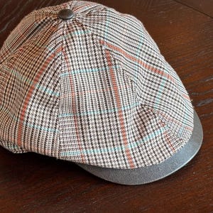 May include: A flat cap featuring a brown, black, and white plaid pattern with thin light blue and orange accent lines. The cap has a rounded crown with a central button at the top and a solid dark gray brim extending from the front edge.