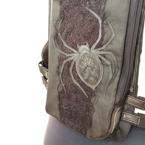 Könnte beinhalten: Schwarze taktische Tasche mit einem großen Spinnen-Applikation. Die Spinne ist in einer helleren Farbe umrandet und hat einen strukturierten Körper. Die Tasche hat einen vertikalen Streifen aus dunklem, strukturiertem Material und einen Reißverschluss.