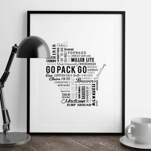 May include: Black and white framed art print featuring a map of Wisconsin filled with text. The text includes phrases like "Go Pack Go", "Brewers", and city names. A black desk lamp and a white coffee cup are in the foreground.