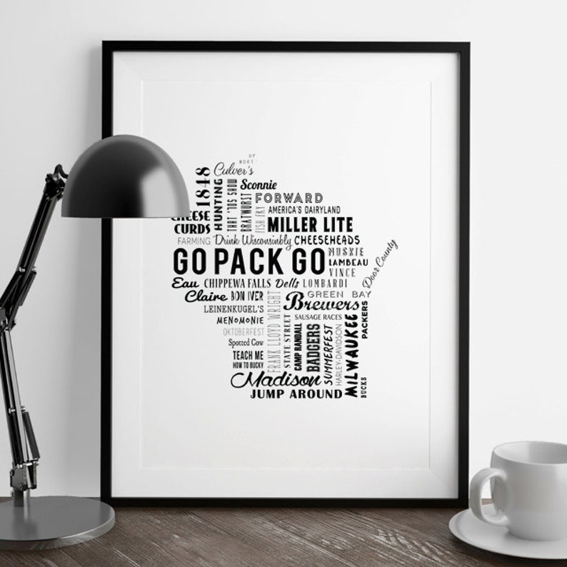 May include: Black and white framed art print featuring a map of Wisconsin filled with text. The text includes phrases like "Go Pack Go", "Brewers", and city names. A black desk lamp and a white coffee cup are in the foreground.