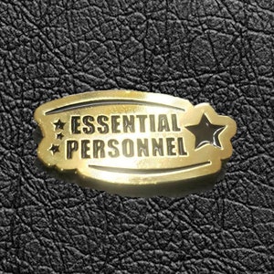 Może przedstawiać: Złota metalowa spinka z czarnym obramowaniem i gwiazdką. Spinka zawiera napis "Essential Personnel" czarnymi literami.