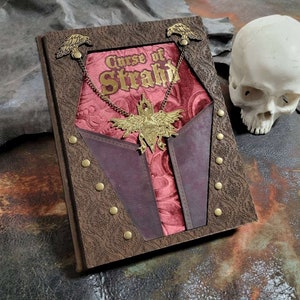 Può includere: Un diario rilegato in pelle marrone con un interno in velluto rosso e un design a forma di pipistrello in metallo dorato. Il diario ha le parole "Curse of Strahd" stampate sulla copertina.
