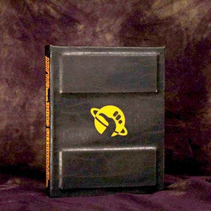 Op de afbeelding: Een zwart hardback boek met een geel en oranje logo van een planeet met een voetafdruk erop. De rug van het boek heeft de tekst "Galaxy Guide - Bomb Seekers".