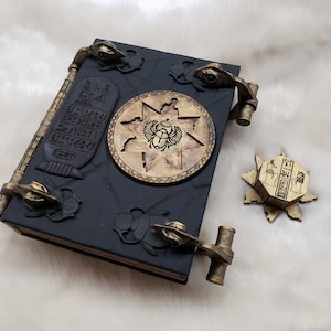 May include: A black leather-bound book with gold accents and intricate carvings. The book has two ornate metal clasps and a circular design featuring an Egyptian scarab beetle. The book is on a white fuzzy surface.