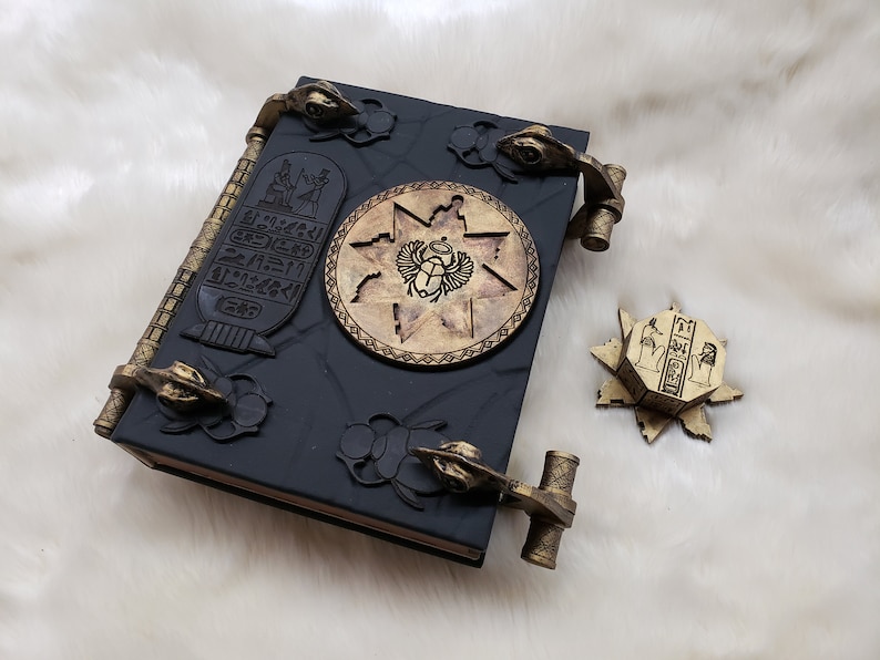 May include: A black leather-bound book with gold accents and intricate carvings. The book has two ornate metal clasps and a circular design featuring an Egyptian scarab beetle. The book is on a white fuzzy surface.