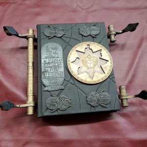 May include: A black leather-bound journal with gold accents and a decorative design featuring scarabs, a star, and an Egyptian hieroglyphic inscription. The journal has two metal clasps with black handles.