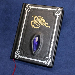 May include: A black leather-bound journal with a silver border and the title "The Dark Crystal" in gold lettering. A purple crystal is embedded in the front cover.