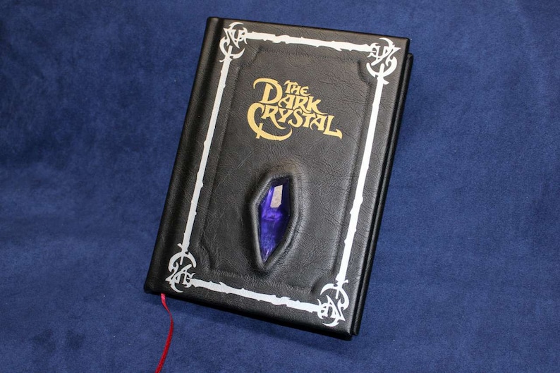 May include: A black leather-bound journal with a silver border and the title "The Dark Crystal" in gold lettering. A purple crystal is embedded in the front cover.