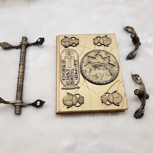 May include: A gold-colored book with an Egyptian theme. The cover features a scarab beetle, an eye of Horus, and hieroglyphics. The book has two ornate gold-colored handles.