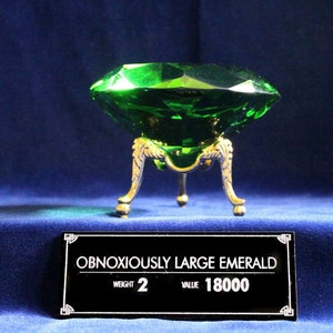 May include: A large, green emerald gemstone on a gold stand. The label reads "Obnoxiously Large Emerald", "Weight 2", and "Value 18000".