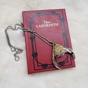May include: A silver necklace with a gold pendant shaped like a stylized claw. The pendant has a detailed design and is resting on a red book with the title "The Labyrinth".