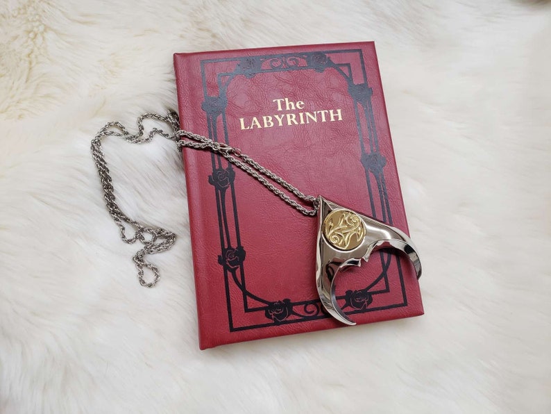 May include: A silver necklace with a gold pendant shaped like a stylized claw. The pendant has a detailed design and is resting on a red book with the title "The Labyrinth".