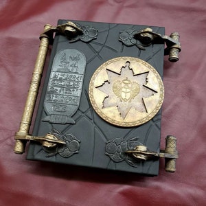May include: A black leather-bound journal with gold metal clasps and a decorative star-shaped design with an Egyptian scarab beetle motif. The journal has a textured surface and a raised design.